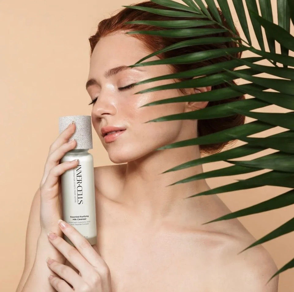 Model holding Inner-Cells Essential Purifying Milk Cleanser against cheek - lifestyle editorial shot with tropical palm leaf background